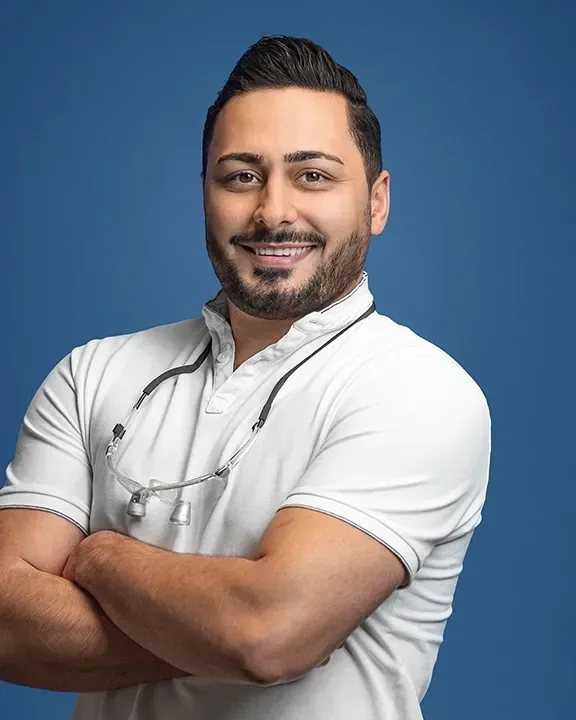 Porträt von Dr. Milad Kazemi im weißen Poloshirt mit verschränkten Armen vor blauem Hintergrund, lächelnd.