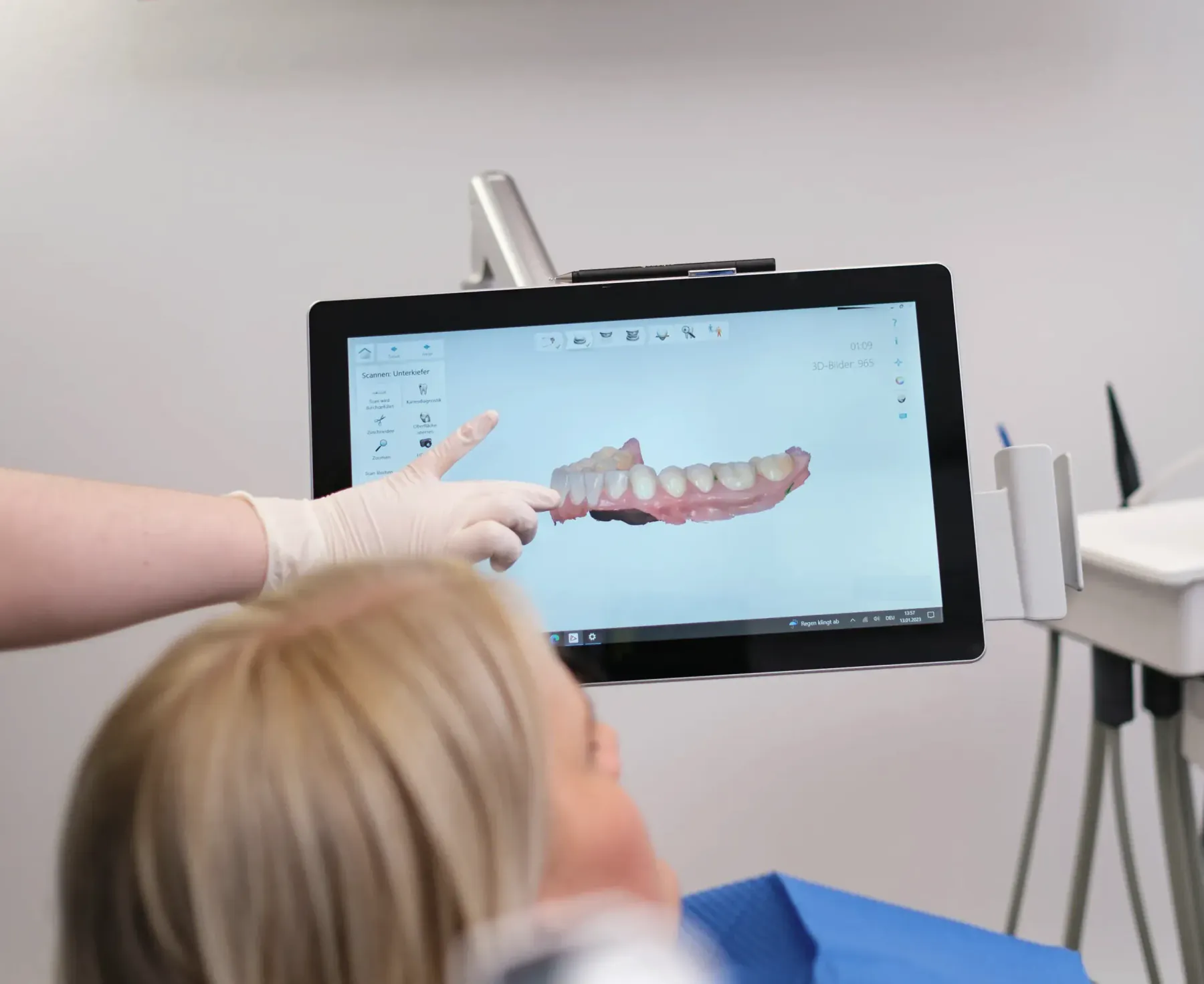 Eine Mitarbeiterin zeigt einer Patientin am Bildschirm den digitalen Scan ihres Unterkiefers. Die Patientin ist von hinten zu sehen.