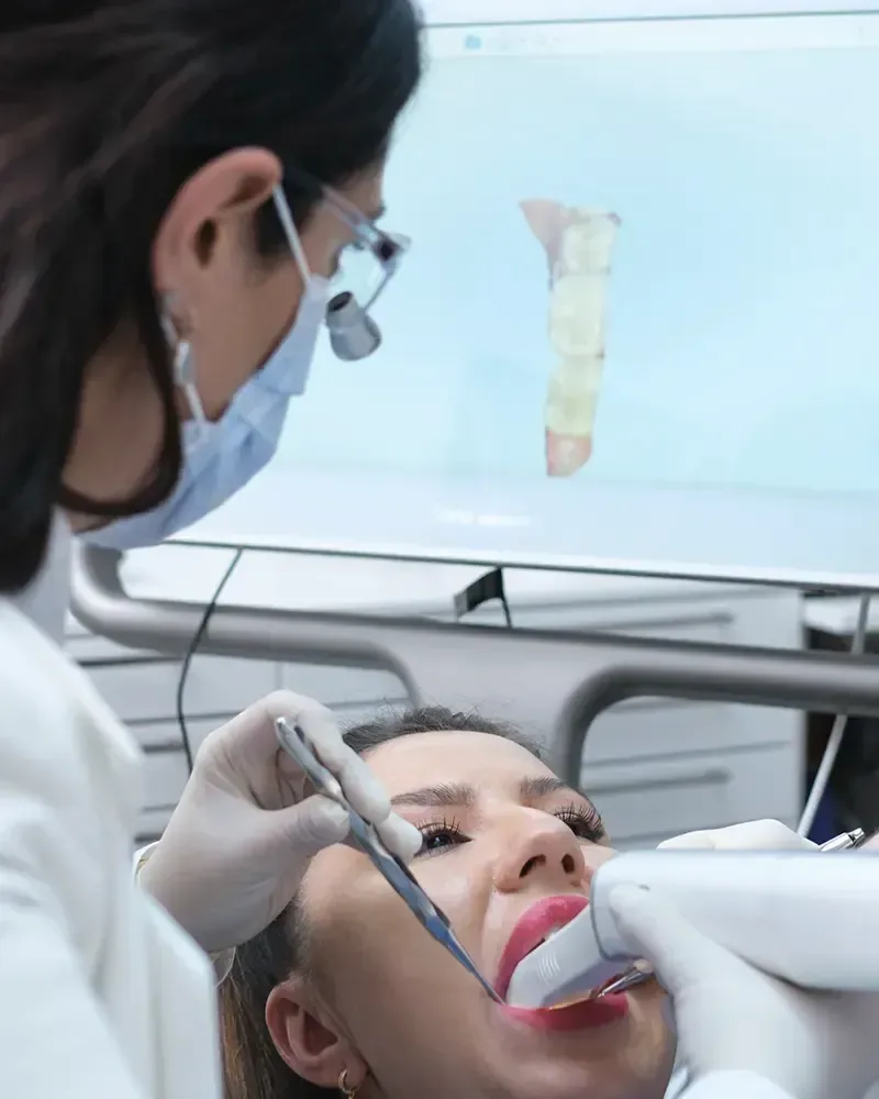 Eine Zahnärztin mit Lupenbrille scannt mit einem Intraoralscanner die Zähne einer Patientin. Im Hintergrund zeigt ein Monitor den Scan.